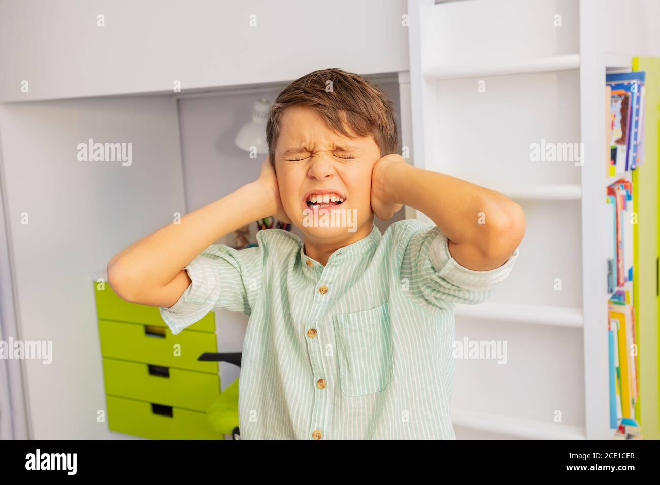 Close portrait of little autistic boy with strong negative face expression closing ears and grins teeth Stock Photo