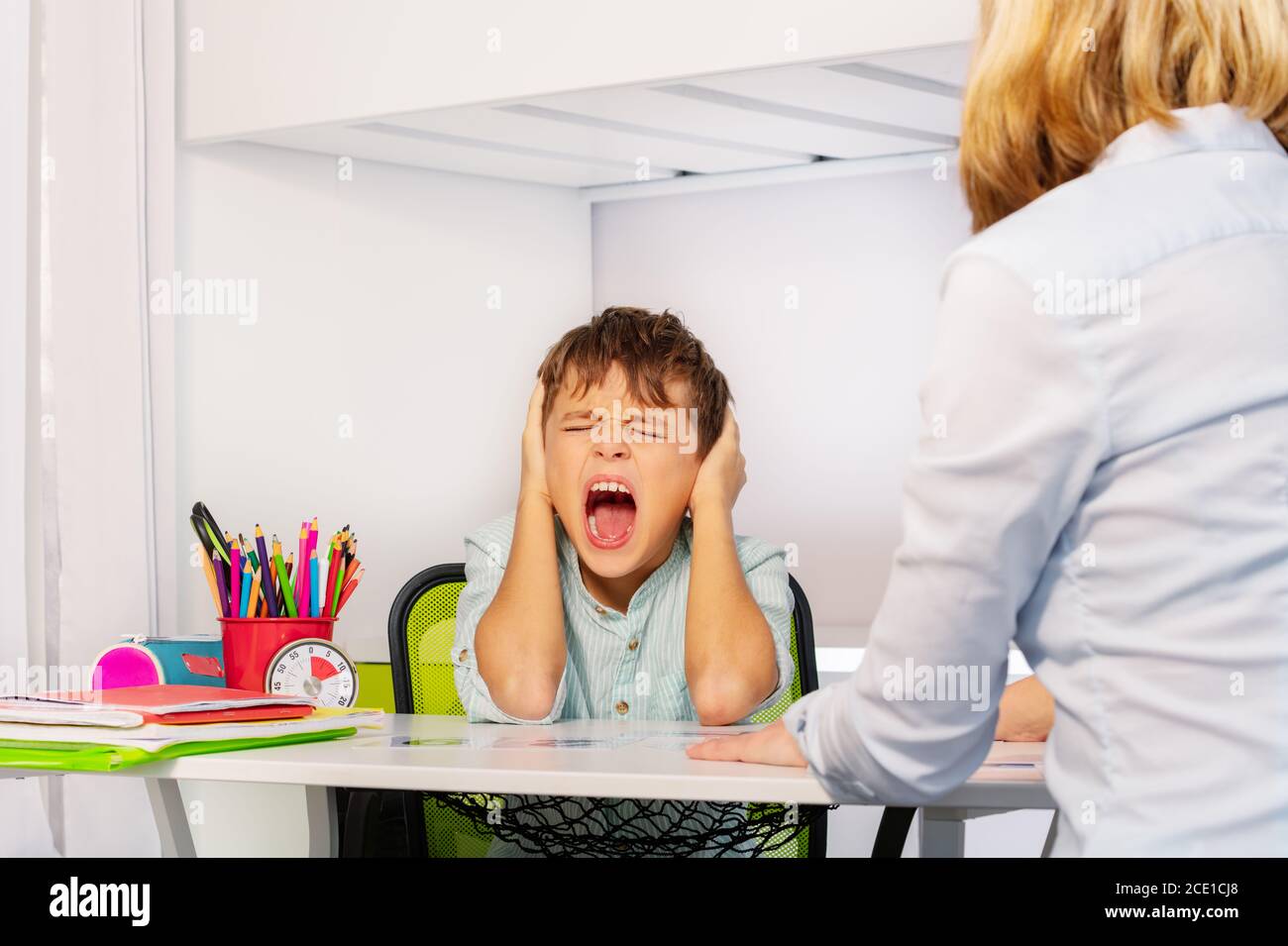 Autistic boy scream and close ears in pain during ABA development therapy sitting by the table with teacher Stock Photo