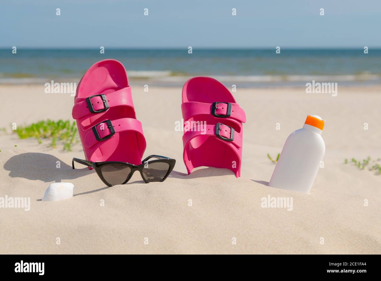 Pink sandals, sunglasses and sunscreen at the beach on a beautiful sunny day. Slippers in the sand by the sea. Flip flops at the shore by the ocean. Stock Photo