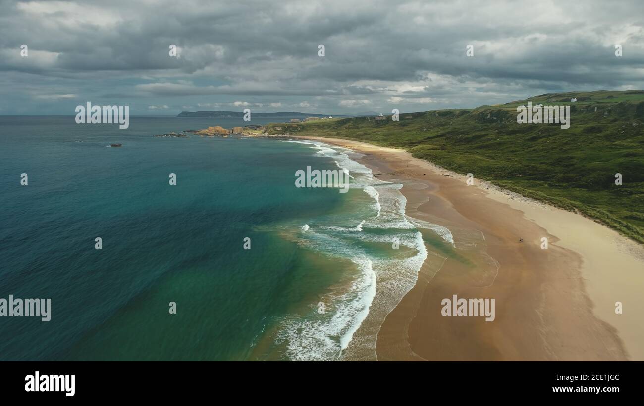 Ireland aerial view: beach ocean bay water crashing. People on shore walk and playing with dogs. Meadows in horizon. Picturesque Northern beauty Stock Photo