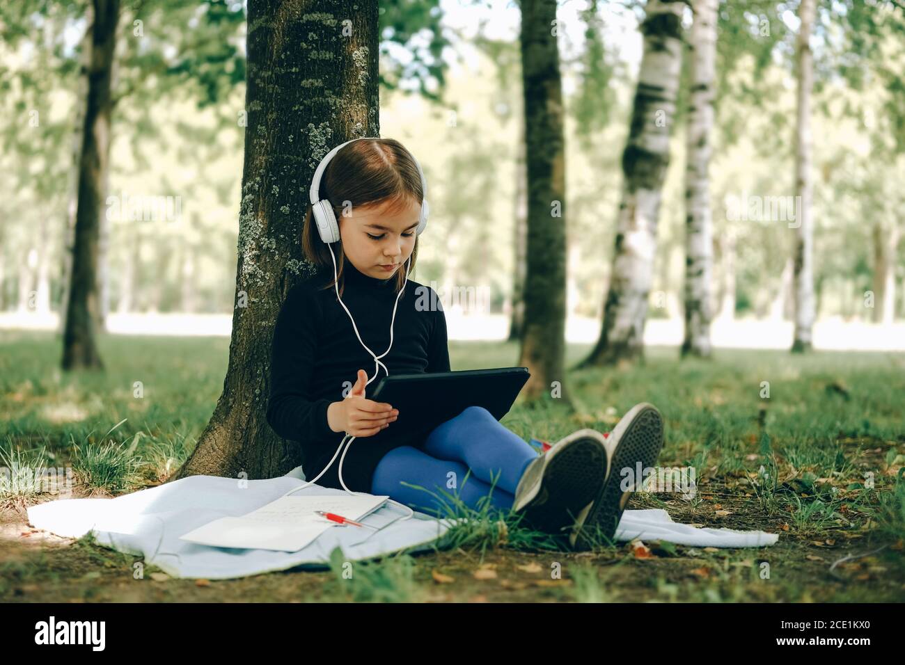 happy girl in white headphones using digital tablet pc in the park. Distant learning concept. Resilience, back to school, new normal. Stock Photo