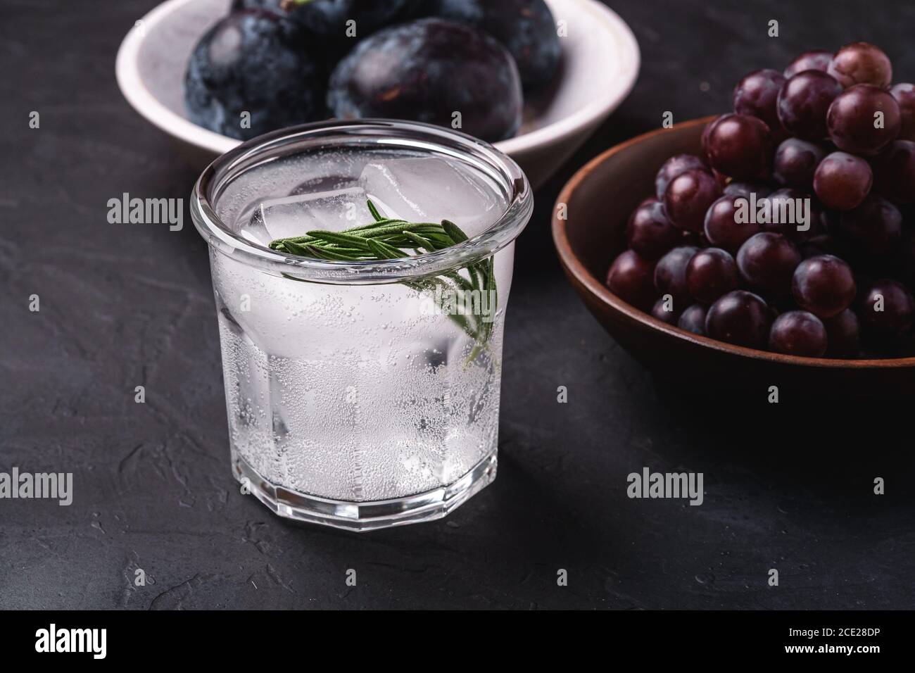 Fresh ice cold carbonated water in glass with rosemary leaf near to wooden bowls with grape and plum fruits, dark stone background, angle view Stock Photo