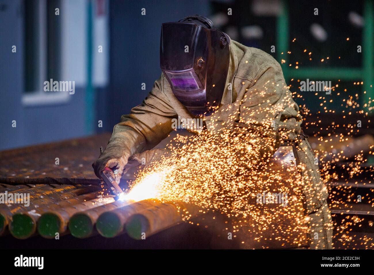Welder in work in factory. Welding steel pipes on manufacturing (producing) plant. Sparks and smoke on dark background. Stock Photo