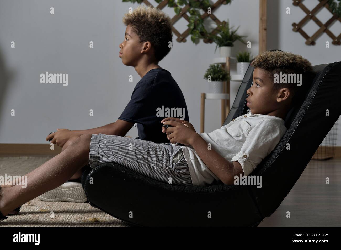 Two mixed race brothers play to play station seated together indoor. Alpha generation offspring addicted with virtual games problem concept Stock Photo