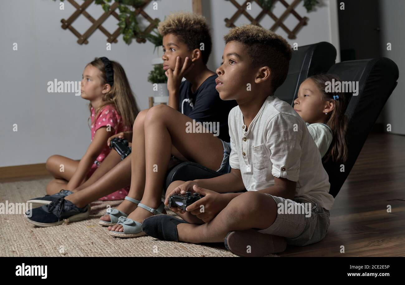 Four pretty multi national kids play to play station seated together at home. Alpha generation offspring addicted with virtual games problem concept Stock Photo
