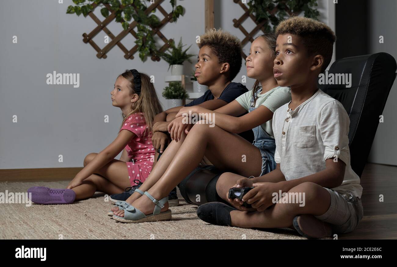 Four pretty multi national kids play to play station seated together at home. Alpha generation offspring addicted with virtual games problem concept Stock Photo