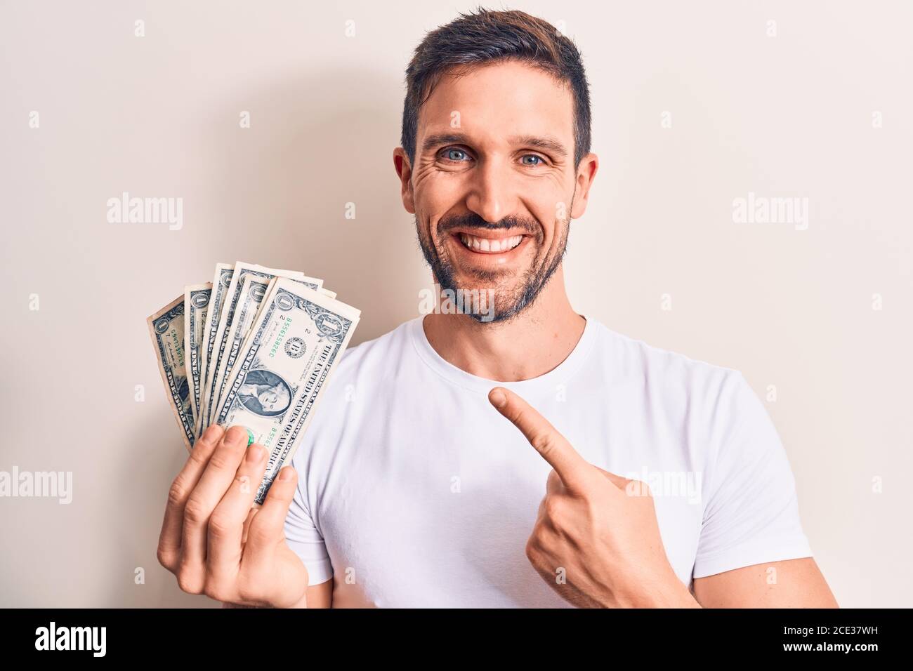 Young handsome man holding bunch of dollars banknotes over isolated white background smiling happy pointing with hand and finger Stock Photo