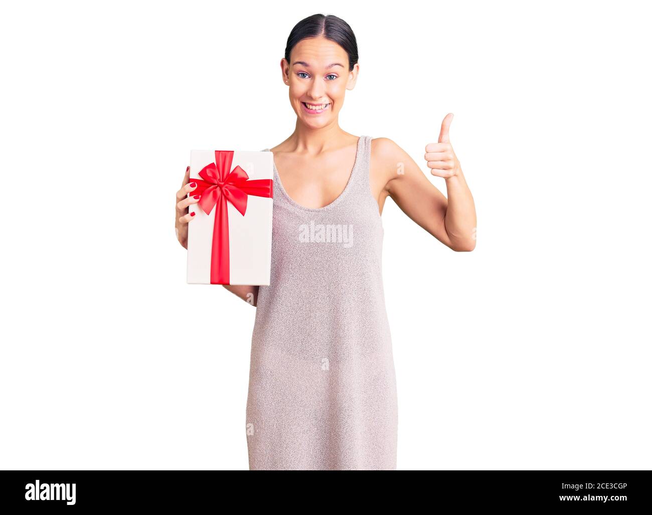 Beautiful brunette young woman holding gift smiling happy and positive, thumb up doing excellent and approval sign Stock Photo