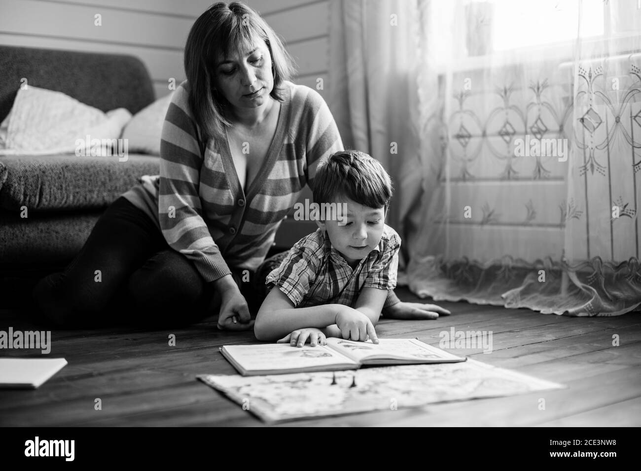 Young woman reading a book with her little son sitting on the floor in home. Black and white photography. Stock Photo