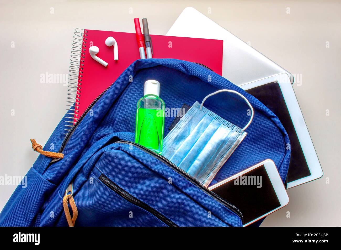 A blue school bag pack with sanitizer and a face mask, pencils earphones, tablet, computer and a smartphone. Back to school during global pandemic. Co Stock Photo