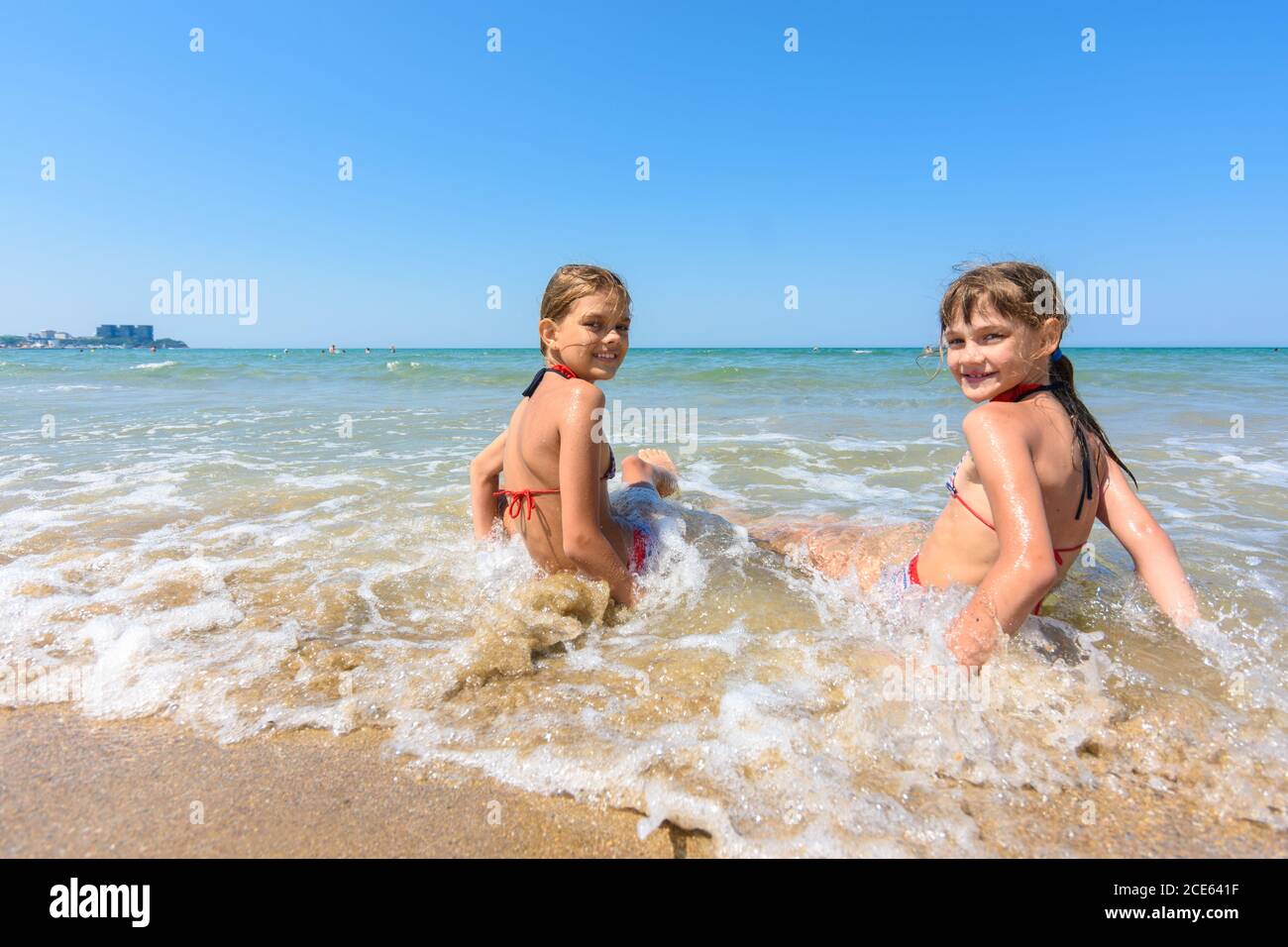 Two girls sit on the sea coast and turn around happily looking at the frame Stock Photo