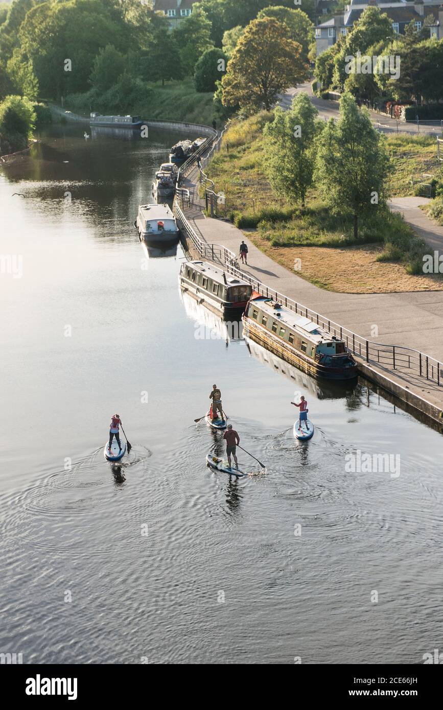Standup paddleboarding (SUP) on the River Avon. Central Bath, UK. Gentle water sport with a group of four friends. River boats moored next to pathway. Stock Photo