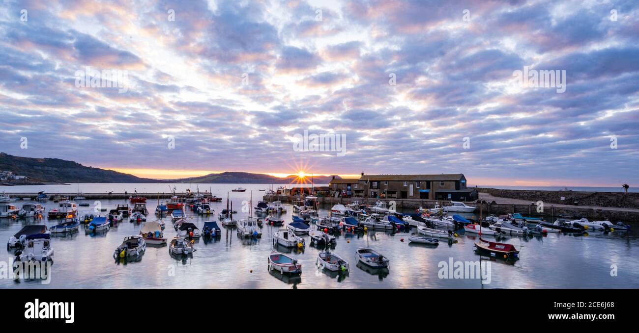 Lyme Regis, Dorset, UK. 31st Aug, 2020. UK Weather: The rising sun bursts through the clouds at sunrise over the Cobb at Lyme Regis on a beautiful, but chilly August Bank Holiday Monday morning. The spectacular clouds in the sky are reflected in the still water of the harbour.  Credit: Celia McMahon/Alamy Live News Stock Photo