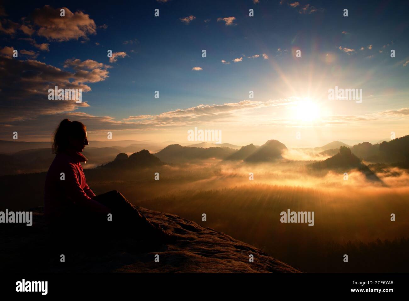 Beautiful  young long hair girl tourist enjoy daybreak on the sharp corner of sandstone rock and watch over valley to Sun. Stock Photo