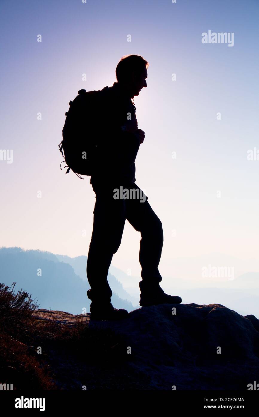 Tourist with sporty backpack stand on rocky view point and watching into misty valley bellow. Sunny misty daybreak in rocky moun Stock Photo