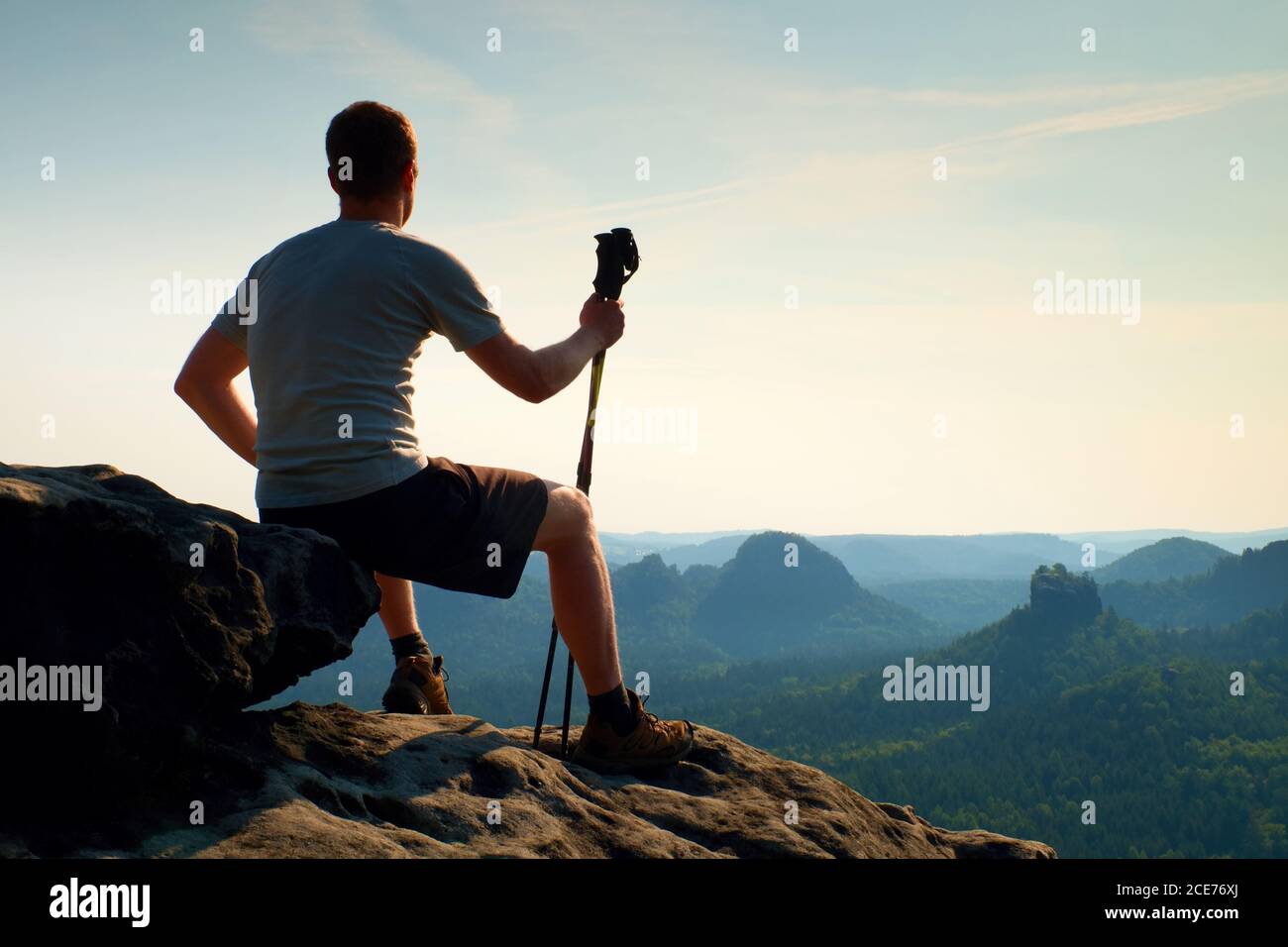 Tired sitting tourist with poles in hand. Sunny spring daybreak in rocky mountains. Hiker with tracking poles sit  on rocky view Stock Photo