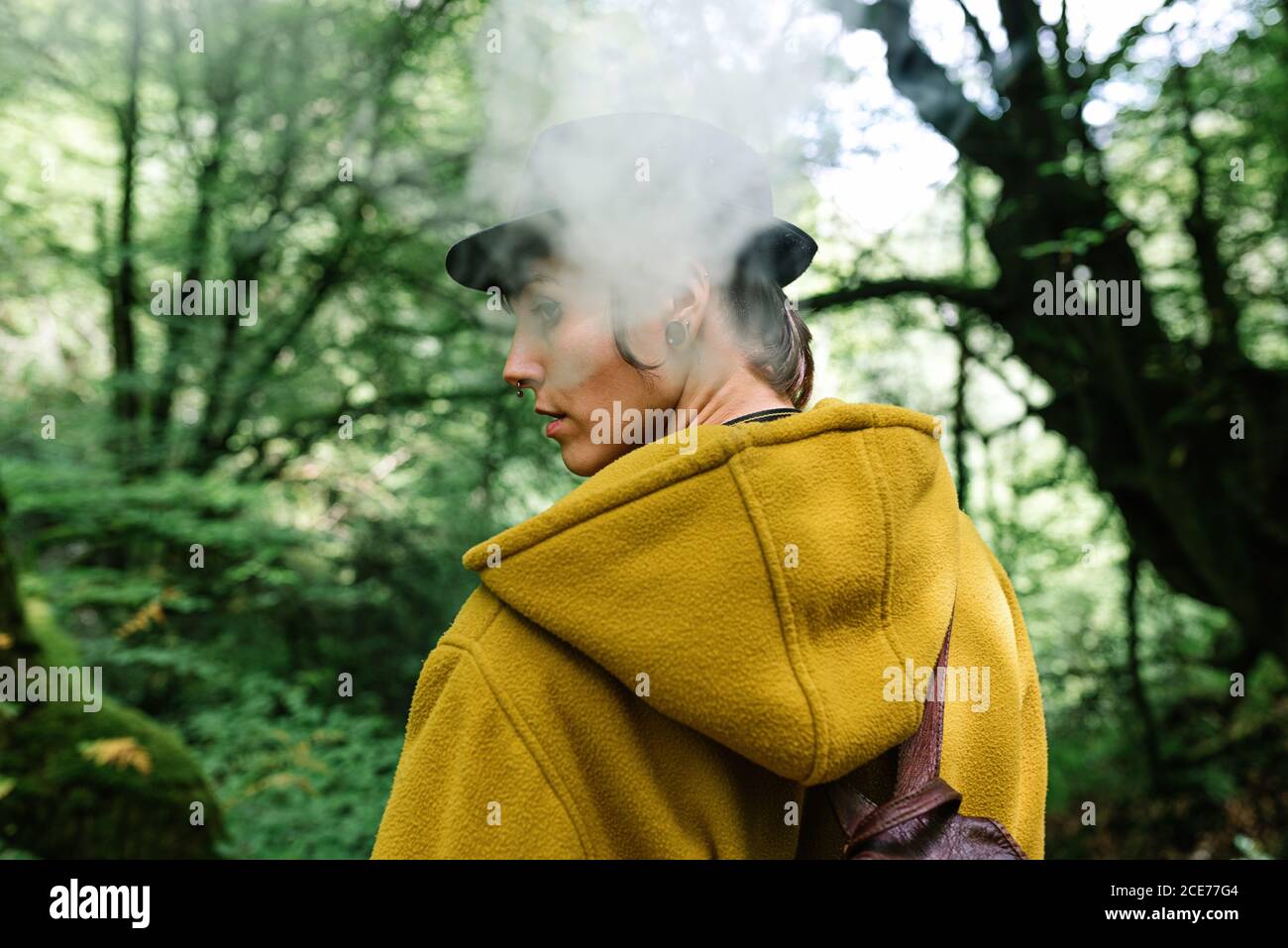 Back view of female traveler with hooded yellow coat exhaling smoke and looking over shoulder on blurred background of green forest Stock Photo
