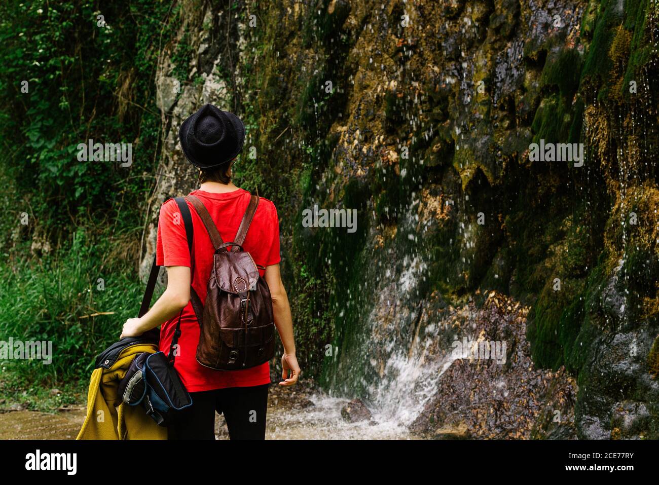 Back view of anonymous tourist in casual clothes and hat standing in woods near wet rock with waterfall and enjoying picturesque scenery Stock Photo
