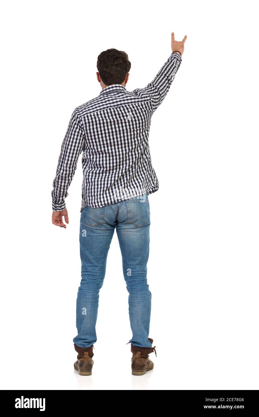 Man in jeans, boots and lumberjack shirt stands with arm raised and shows sigh of the horns. Rear view. Full length studio shot isolated on white. Stock Photo