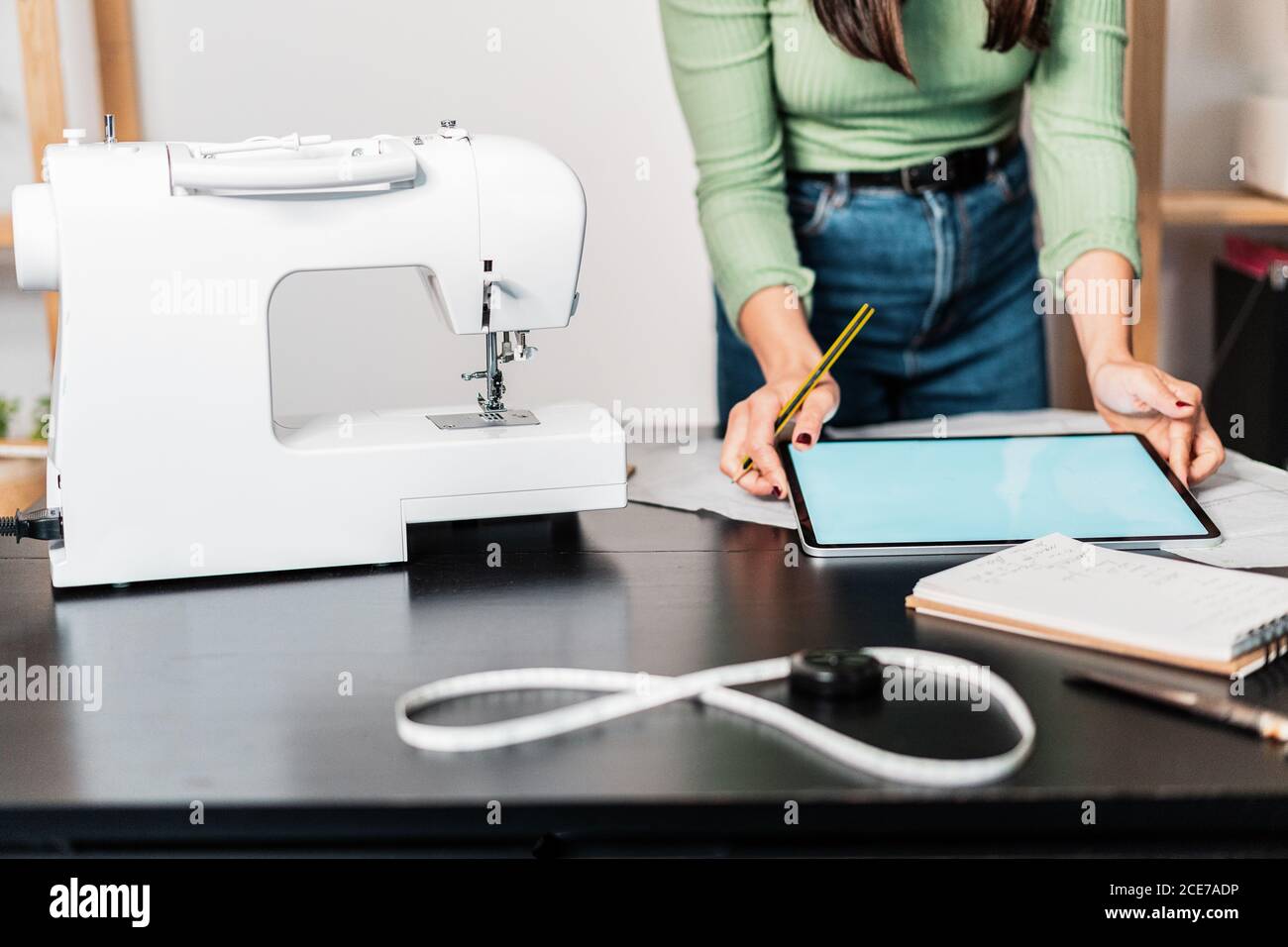 Crop unrecognizable seamstress standing leaned forward while using tablet and straightedge at table with papers in studio Stock Photo