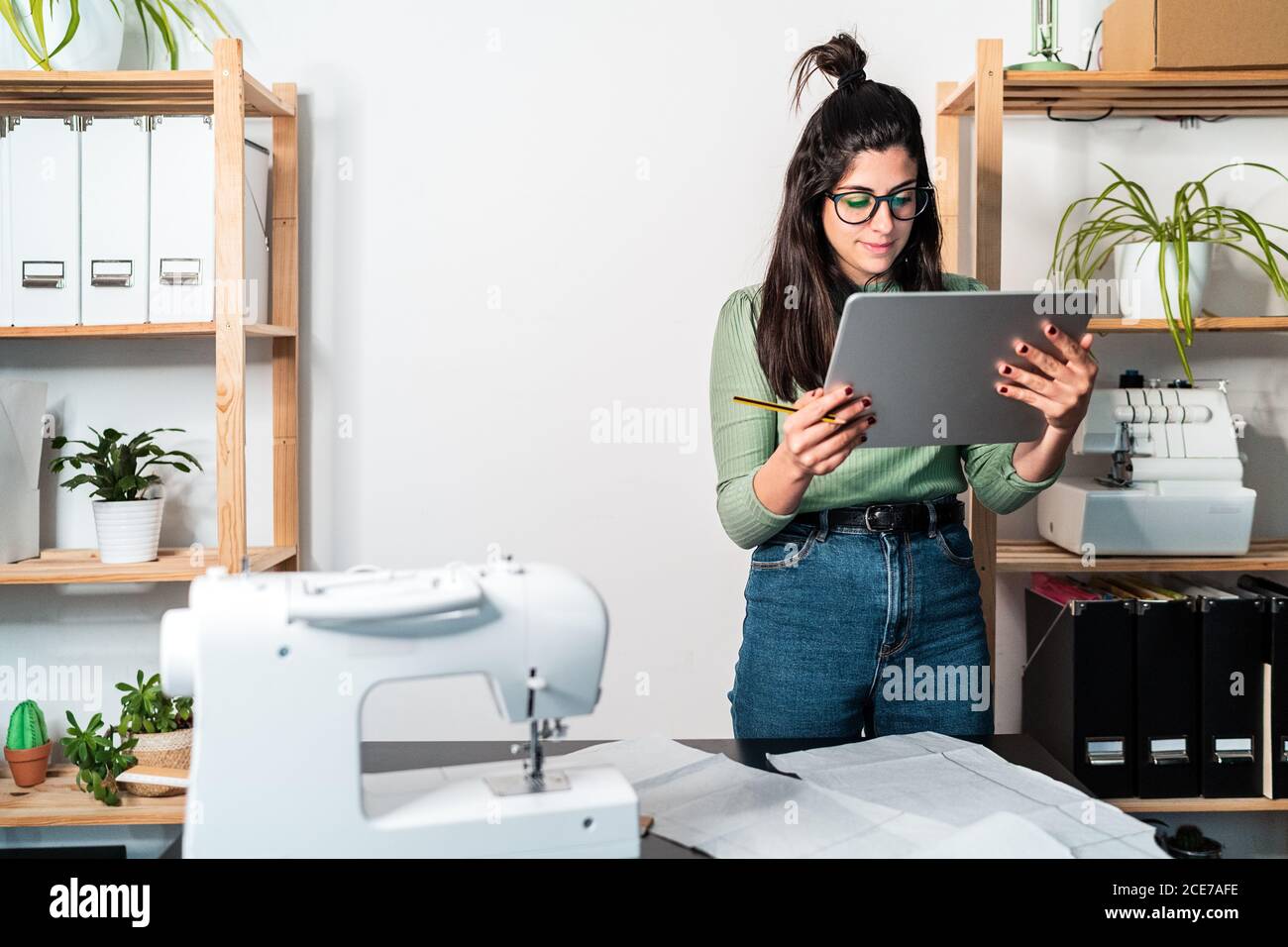 seamstress standing leaned forward while using tablet and straightedge at table with papers in studio Stock Photo