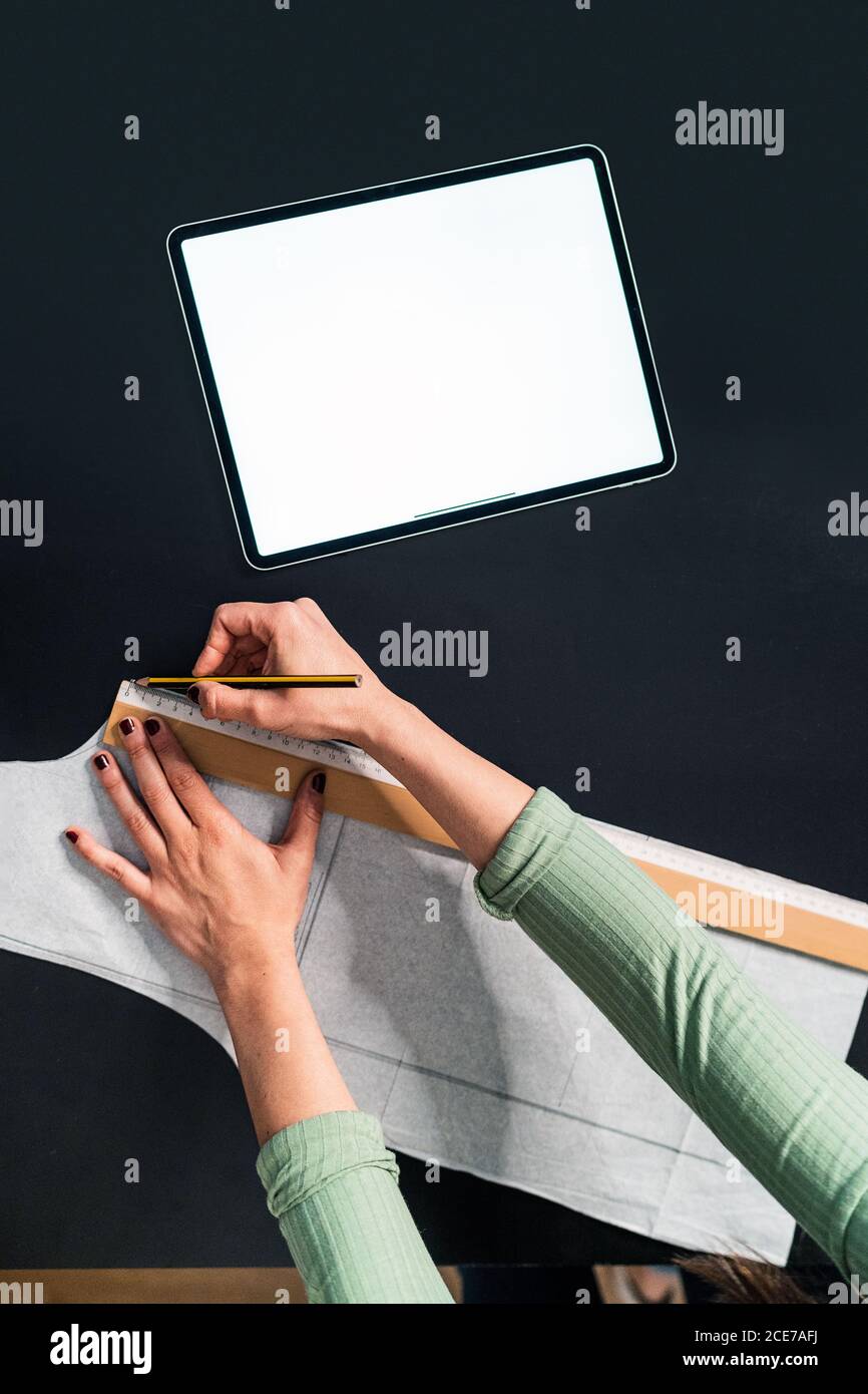 from above crop seamstress standing leaned forward while using straightedge at table with papers and tablet in studio Stock Photo