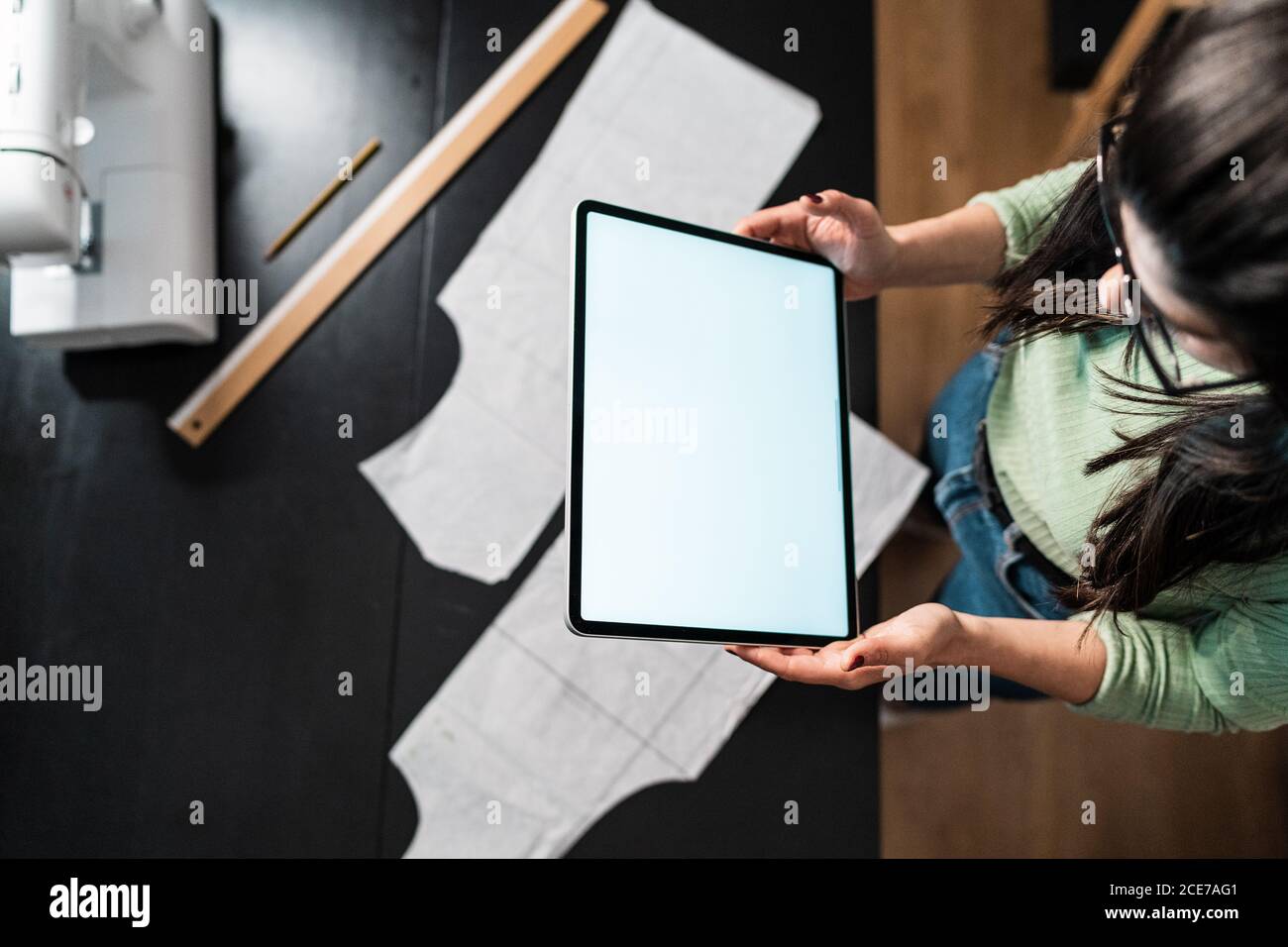 from above crop seamstress standing leaned forward while using tablet and straightedge at table with papers in studio Stock Photo