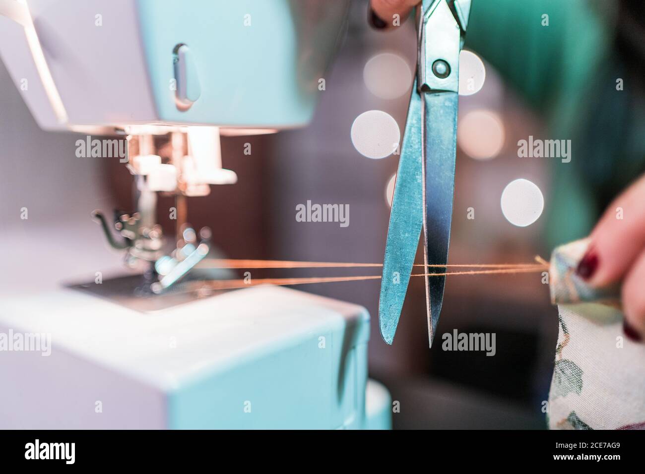 Crop unrecognizable craftsWoman extending thin thread while cutting with scissors on modern metal sewing machine on table in workshop Stock Photo