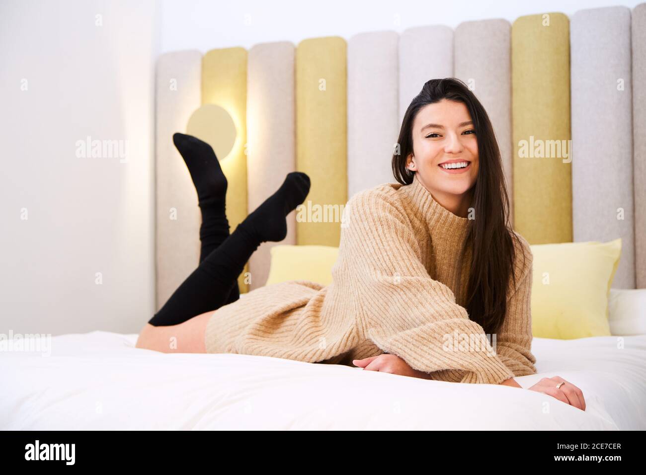 Charming female in oversize sweater and stockings lying on soft bed in modern bedroom and cheerfully looking at camera Stock Photo