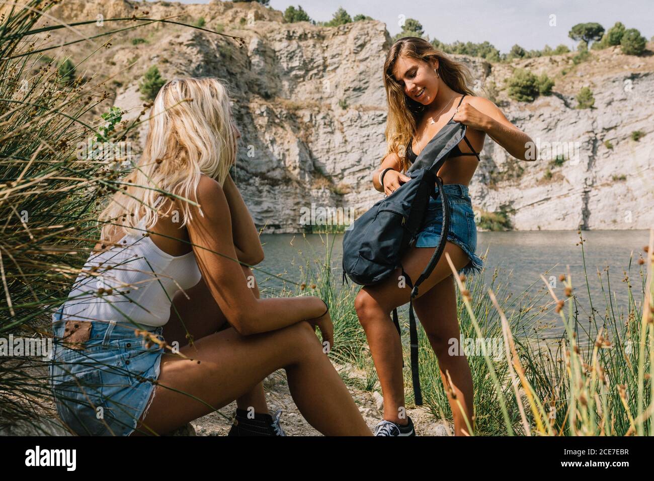 Content female tourists relaxing together on stony shore near lake during summer vacation Stock Photo