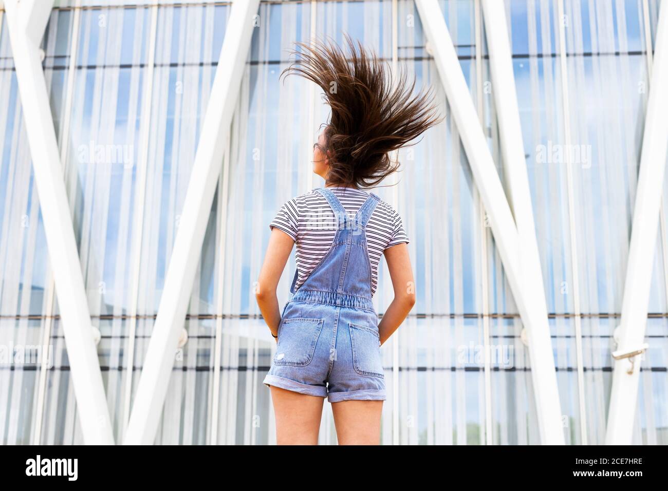 Back view of unrecognizable teen girl in casual outfit shaking long hair near modern building on city street Stock Photo