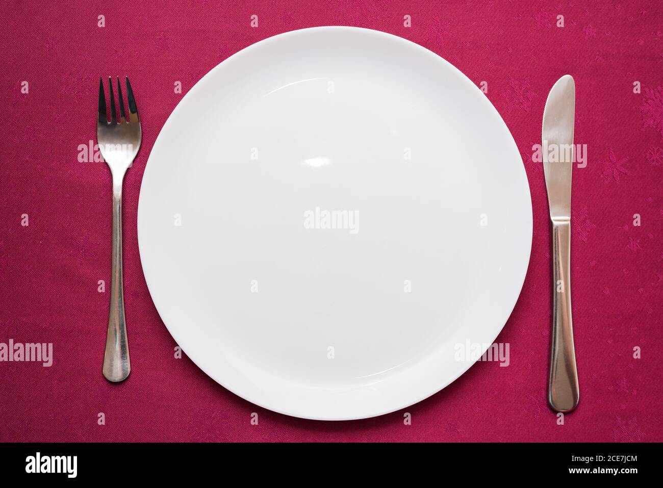 Empty white plate, fork and knife on maroon tablecloth. Elegant table setting. Top view, close-up Stock Photo