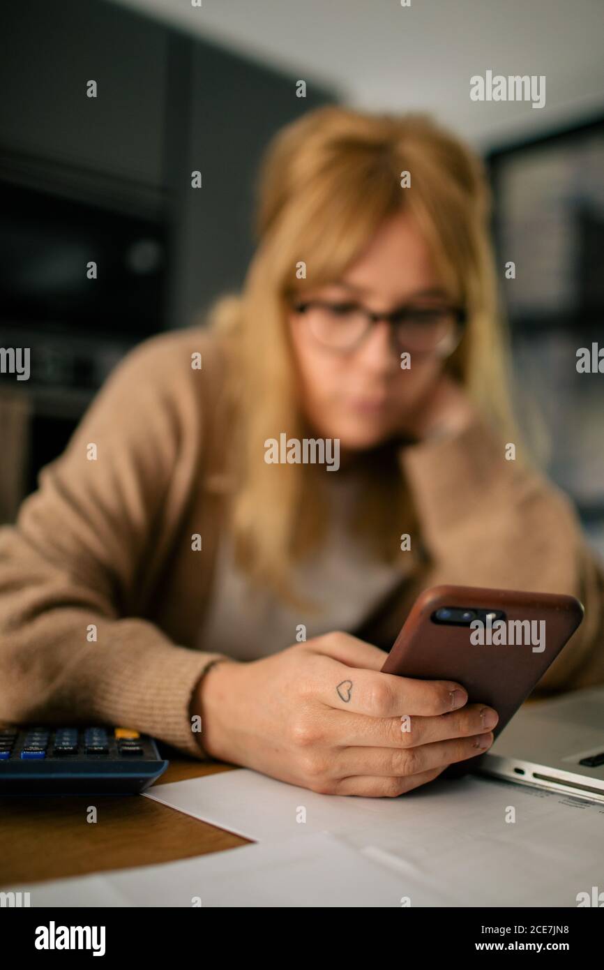 Side view of pensive female accountant sitting at table using on mobile phone while having break during remote work at home and laughing Stock Photo