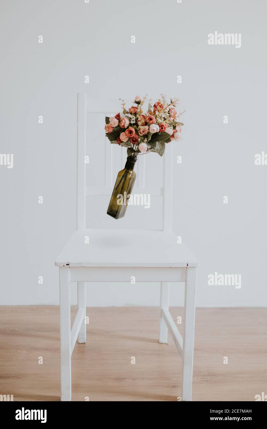 Vintage bridal flower bouquet placed inside a glass vase floating on air over a white wooden chair in an empty room with white walls Stock Photo