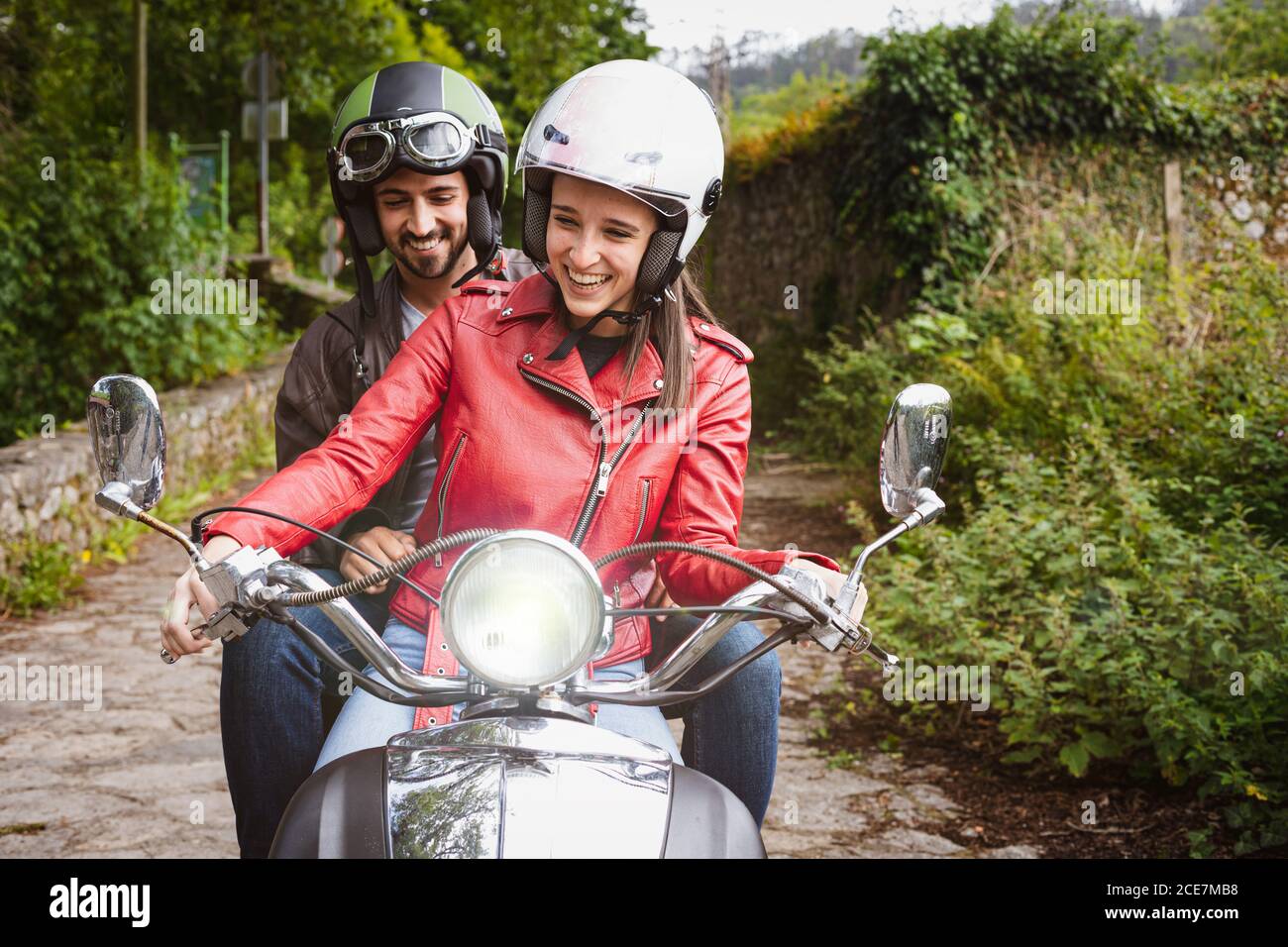 Cheerful man in helmet sitting on motorcycle with smiling girlfriend in leather jacket riding on pathway near lush green shrubs on weekend Stock Photo
