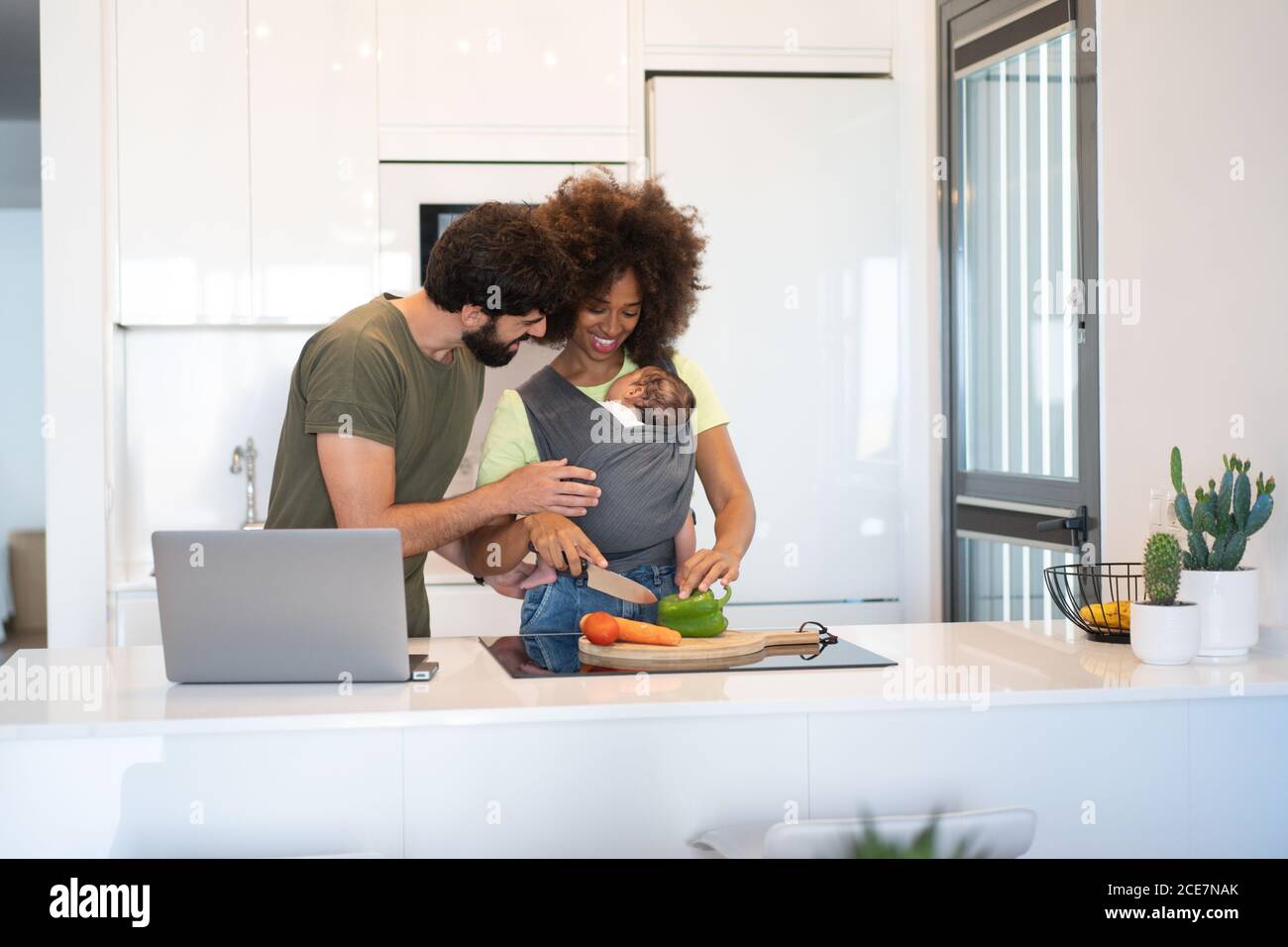 Content black mother with baby in sling cutting vegetables and Arab father working on laptop while standing at counter in kitchen Stock Photo