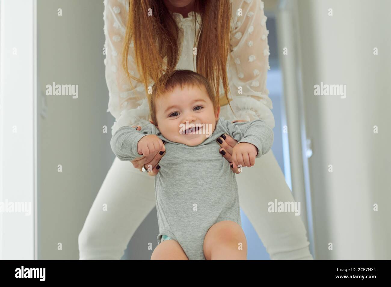 Cute cheerful toddler in gray bodysuit held by crop unrecognizable mother smiling and looking at camera  while having fun together at home Stock Photo