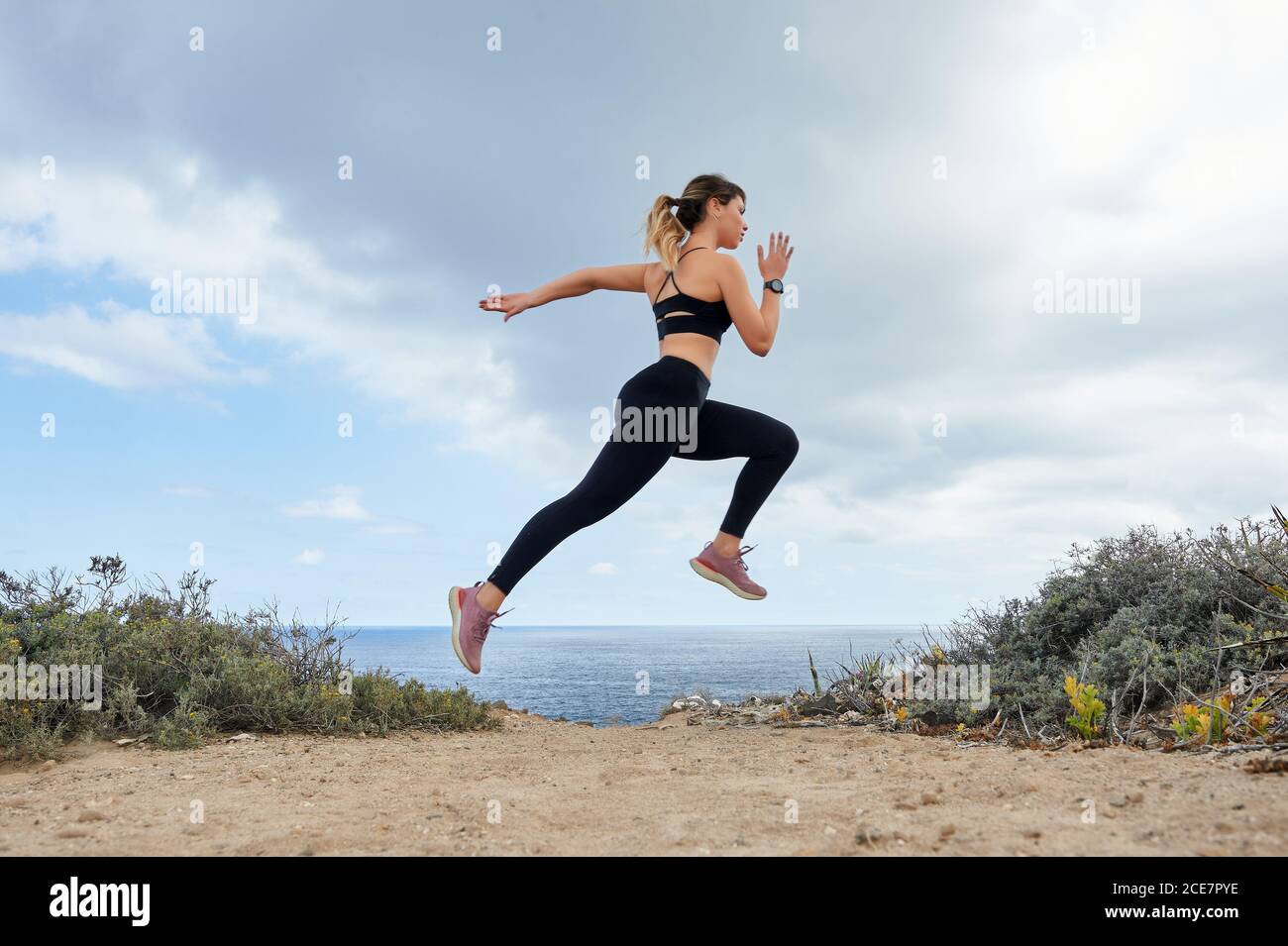 Side view full body slim determined female runner in black leggings and sports bra jogging and jumping on seashore against calm blue sea during sunny day Stock Photo