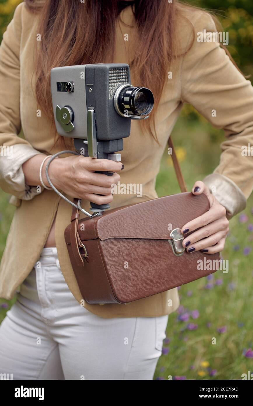 Crop unrecognizable slim female photographer in jacket standing on grass with leather case and retro video camera in countryside in summer Stock Photo