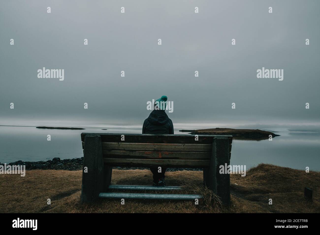 Back view of anonymous tourist in warm clothes and hat relaxing on wooden bench and admiring ocean view from hill on overcast day in Iceland Stock Photo