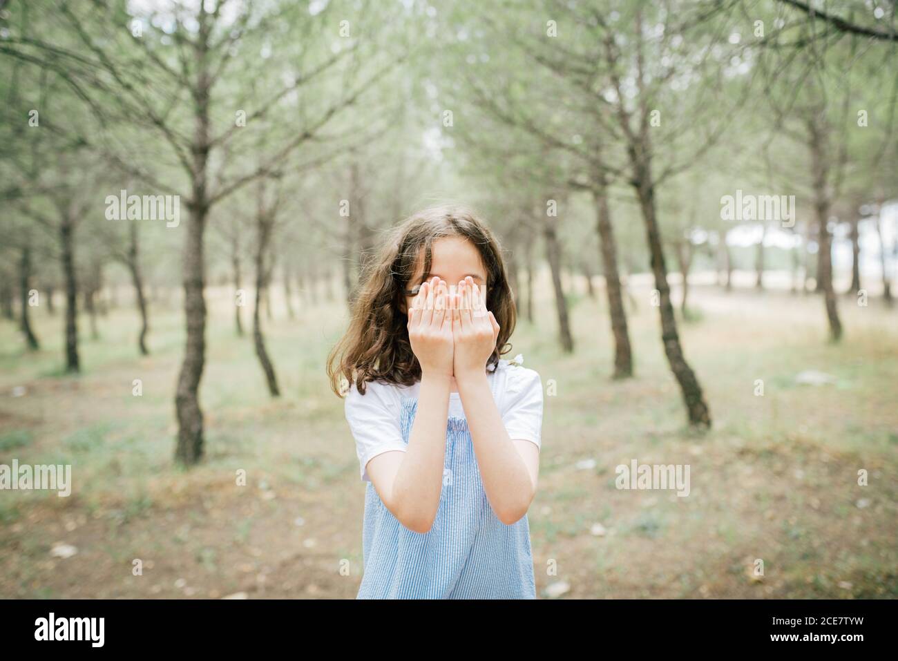 Full body of slim barefoot child in casual dress standing on chair covering face with hands in city park in daylight Stock Photo