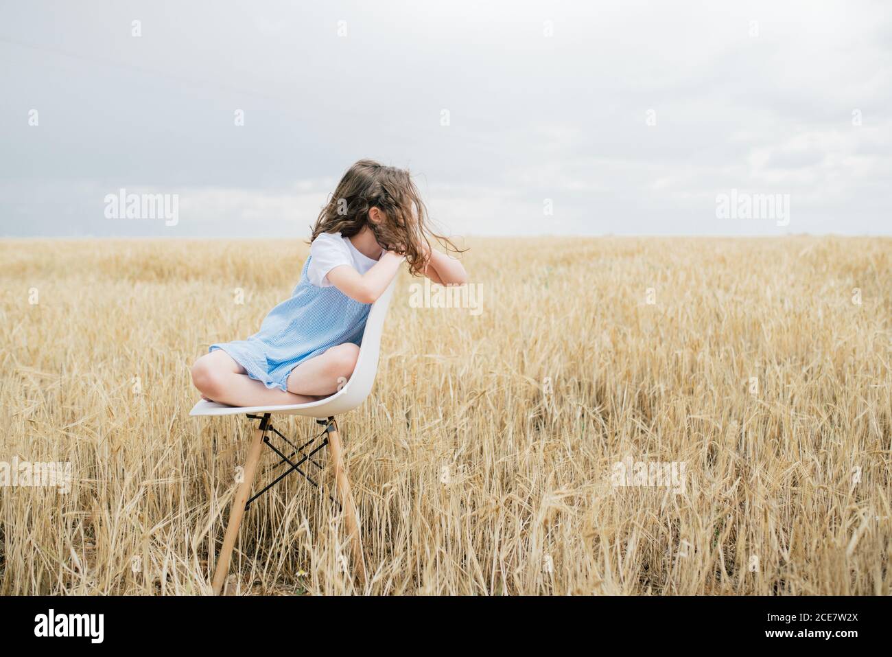 Alone ponder barefoot child with brown hair sitting on chair embracing knees in field with faded grass in overcast weather in autumn and looking away Stock Photo