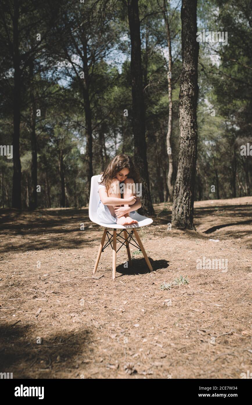 Lonely barefoot girl in dress sitting on stool embracing knees and looking down in forest with high trees under blue sky in summer Stock Photo