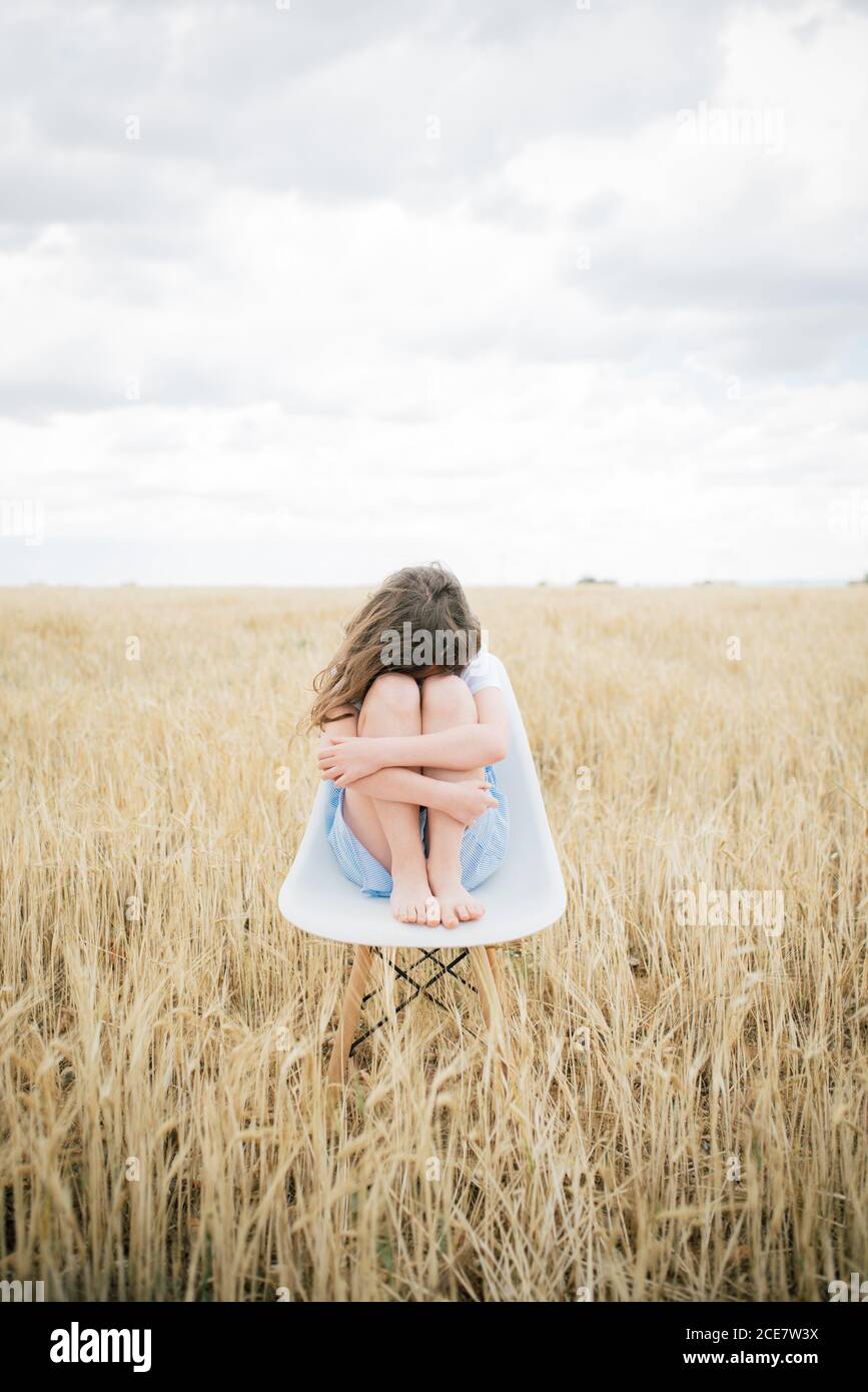 Alone ponder barefoot child with brown hair sitting on chair embracing knees in field with faded grass in overcast weather in autumn covering face Stock Photo
