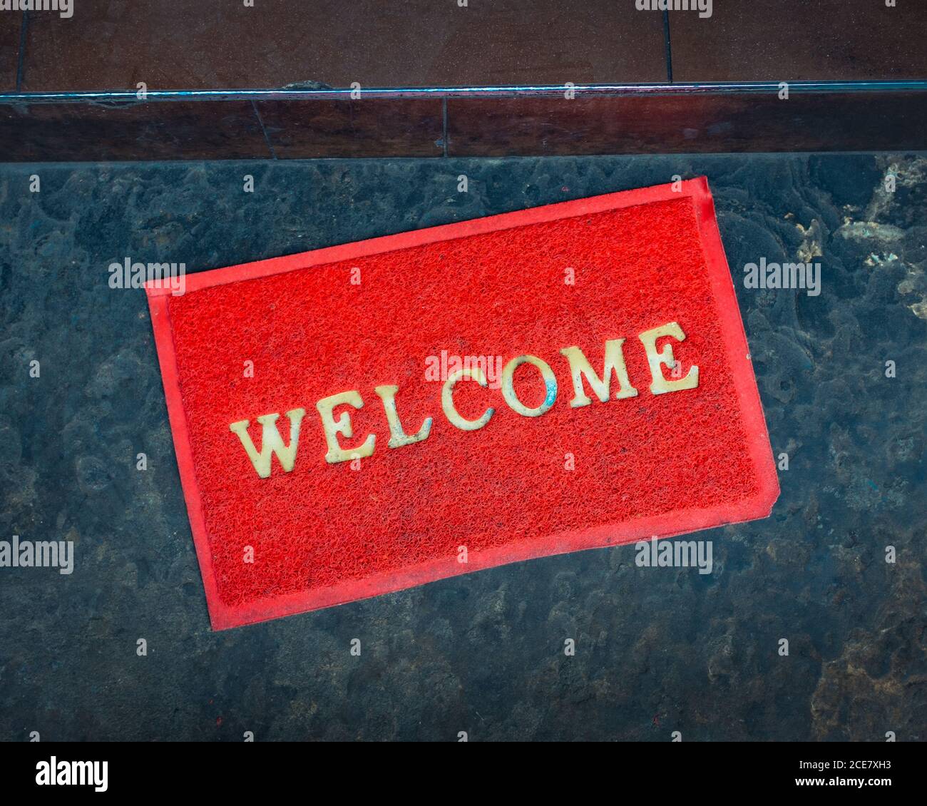 From above rectangular red doormat with word welcome of golden color placed on dark blue marble floor near apartment entrance Stock Photo