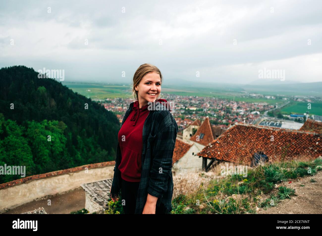 Cheerful female in outerwear standing on high viewpoint looking at camera scenery of old town on overcast day in Saint George, Transylvania, Romania Stock Photo