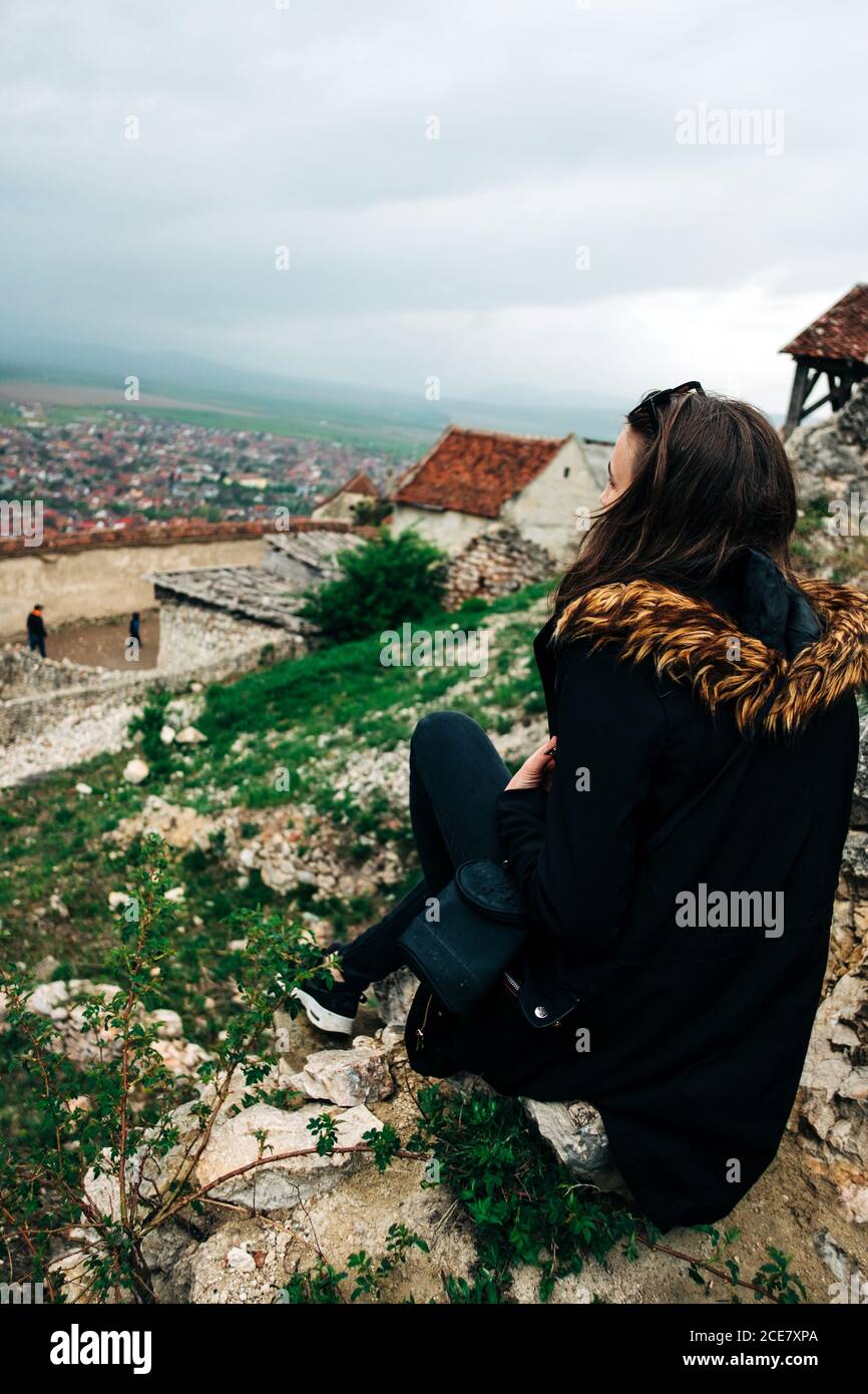 Side view full body unrecognizable calm female in outerwear sitting on high viewpoint and admiring amazing scenery of old town on overcast day in Saint George, Transylvania, Romania Stock Photo