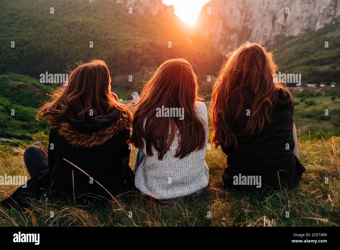 Back view of anonymous female friends chilling on grass in mountains during picturesque sunset in Transylvania and looking over shoulder Stock Photo
