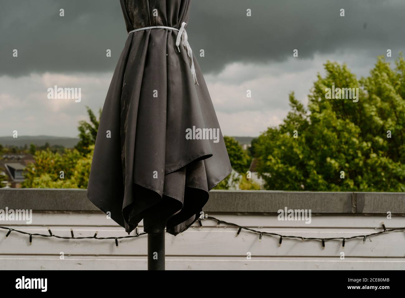 Closed black garden umbrella on terrace against leaden dark cloudy sky before heavy rain on overcast summer day Stock Photo