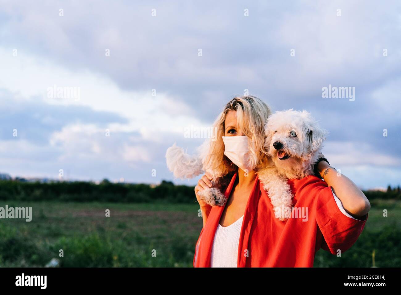 Positive female in casual clothes wearing protective face mask standing with funny white Bichon Frise dog on shoulders against blurred summer countryside landscape during coronavirus outbreak Stock Photo